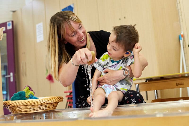 A woman sits her young child on her knee, they are next too a tub of water and the child's legs are being playfully splashed with water.