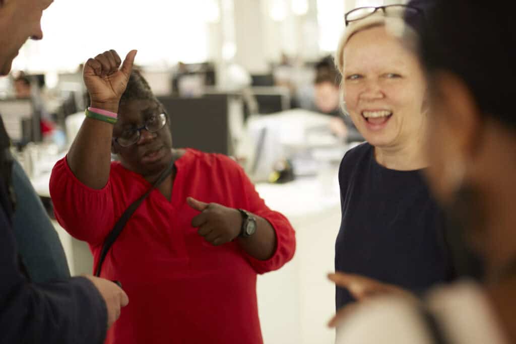 Two people are communicating in sign language whilst other people watch and smile. 