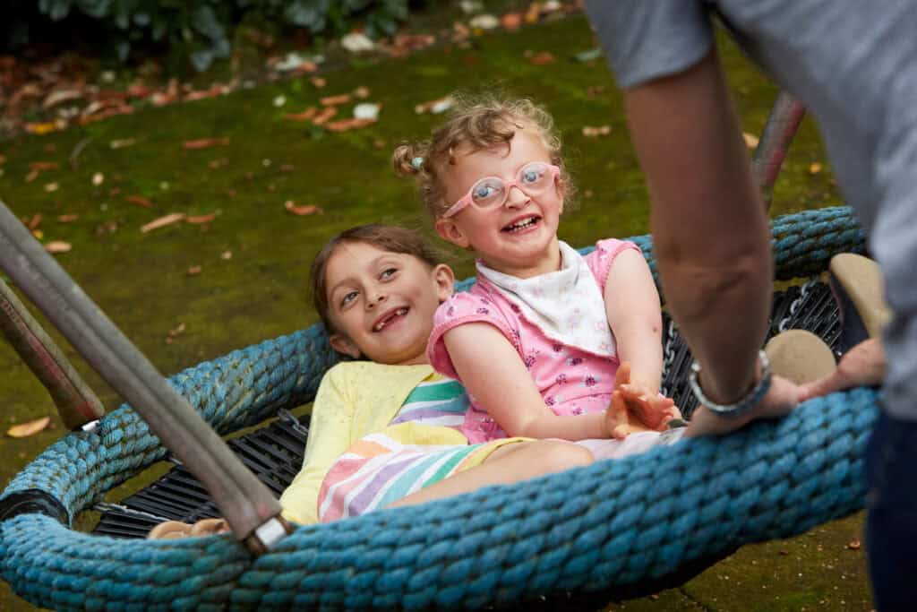 Two young girls, one wearing pink glasses, are swinging together on a swing outdoors.