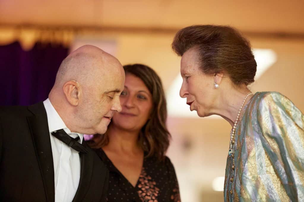 HRH The Princess Royal meets and older gentleman with a shaved head wearing a tuxedo. She leans in close to speak to him. Her hair is darn brown and styled up and she is wearing a metallic gold and silver jacket.