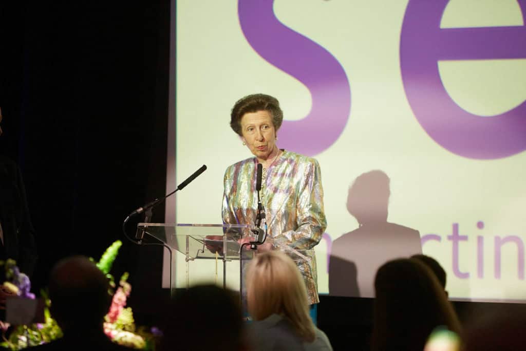 HRH The Princess Royal stands on a stage giving a speech. Her hair is dark brown and styled up. She is wearing a metallic patterned jacket and behind her we see the Sense logo projected onto a large screen.
