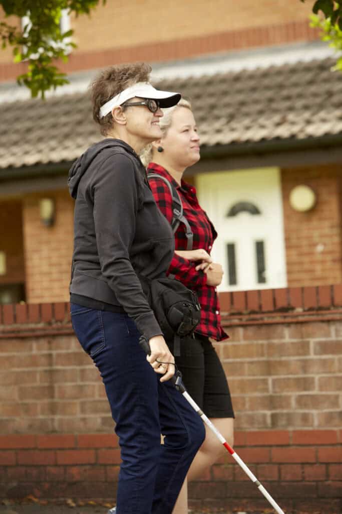 A woman with short hair and a white visor and cane being guided by another woman.