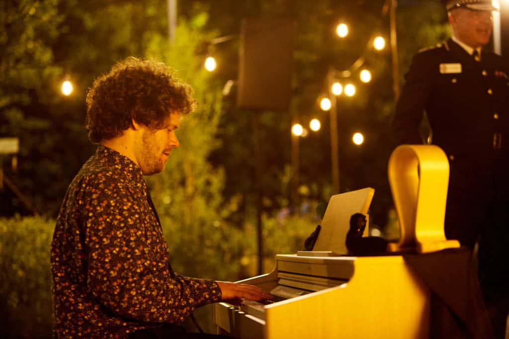 A man with brown curly hair wearing a floral shirt plays the yellow piano outside in the garden. A man in full police uniform stands to the right of the piano.