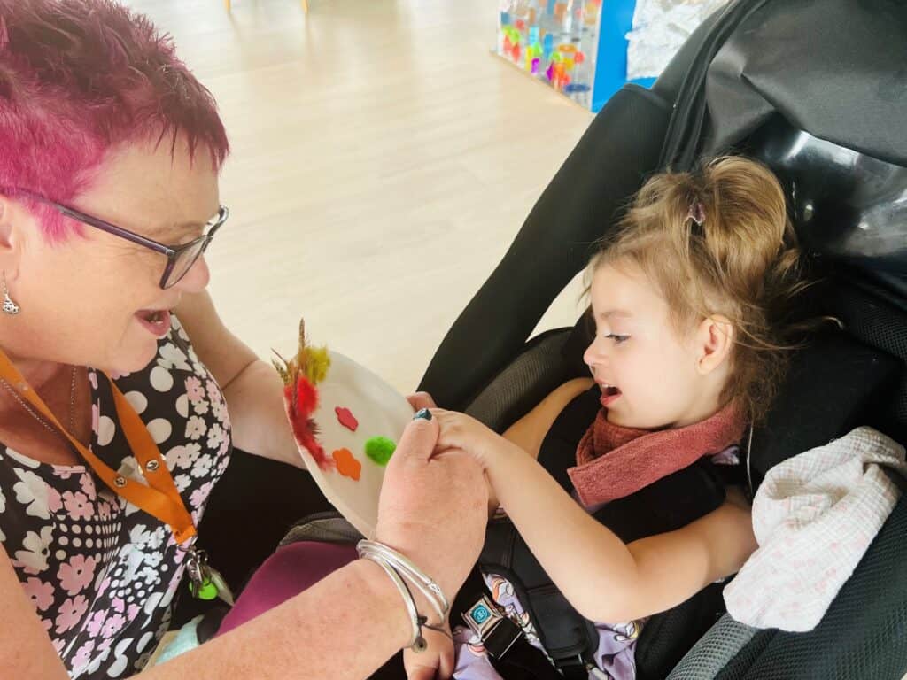 Anne, a support worker with short pink hair, playing with Verity, a small girl in a pushchair.
