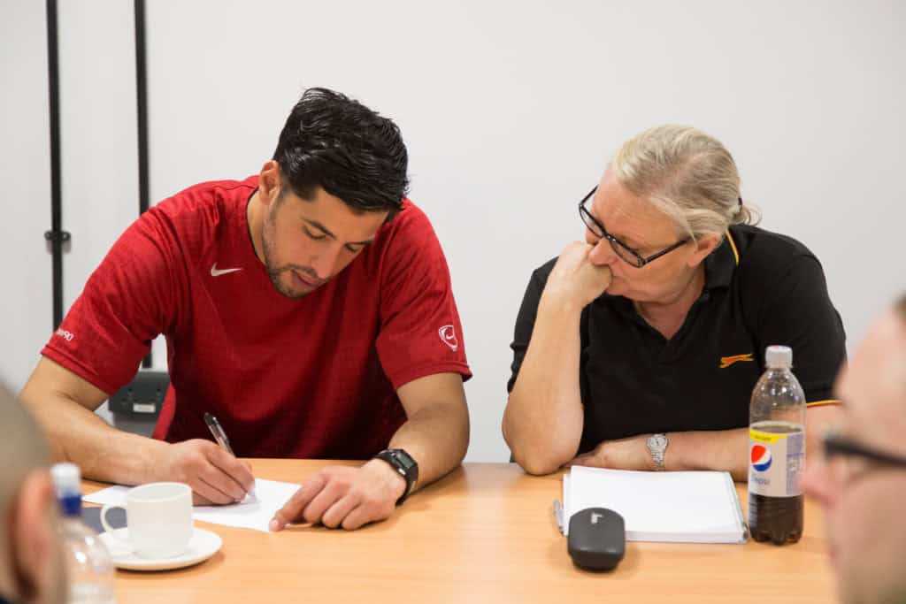 Two people sat at a table, one is writing notes on paper whilst the other looks on