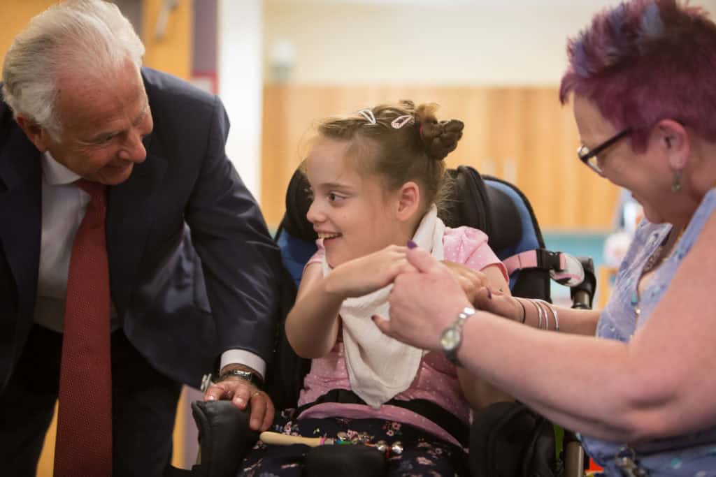 The Lord Levy standing next to a young blonde girl in a wheelchair and a woman with purple hair.