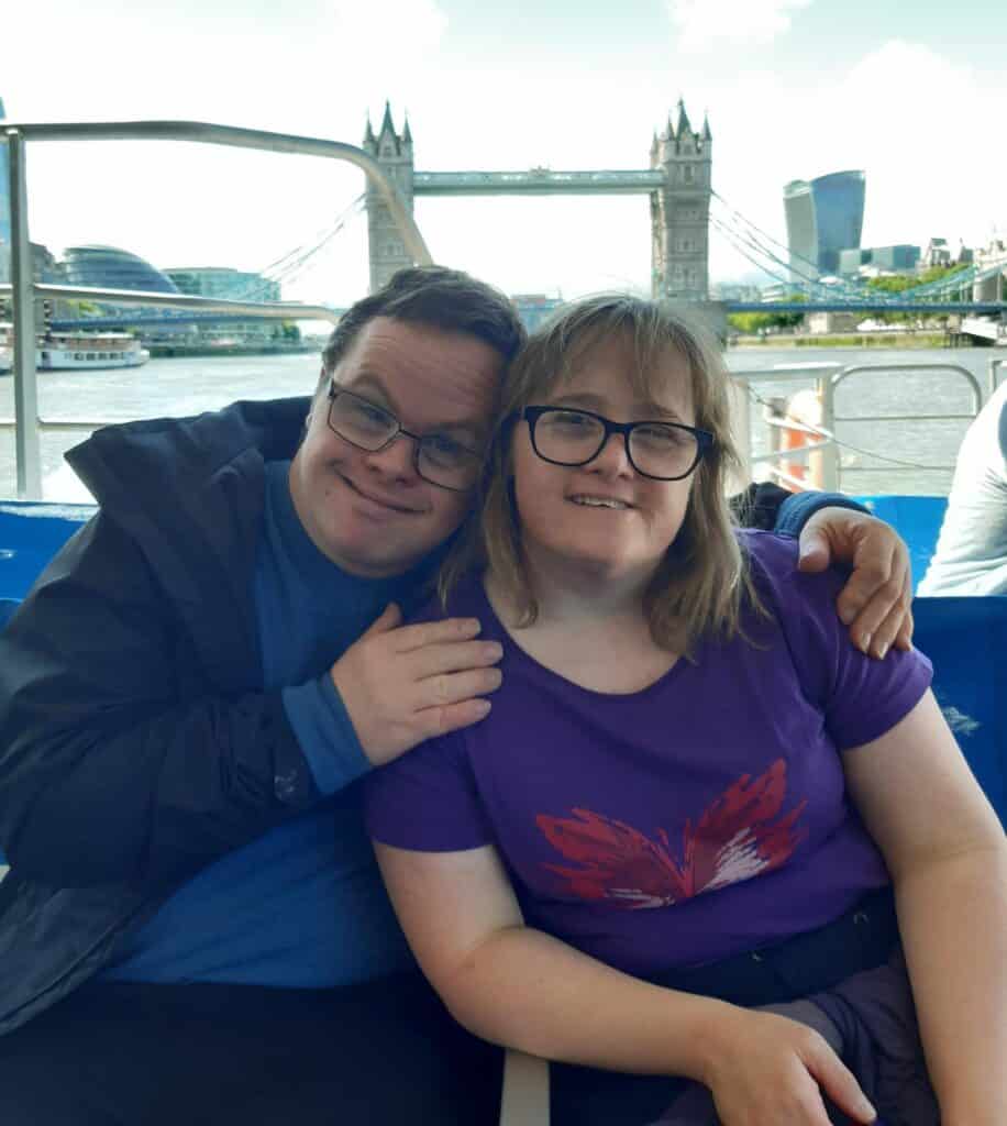 A young man sits with his arms around his girlfriend. Behind them is the River Thames and Tower Bridge.