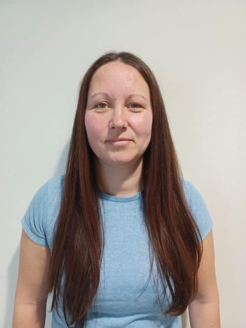 A headshot of a woman with long dark brown hair, wearing a pale blue t-shirt.