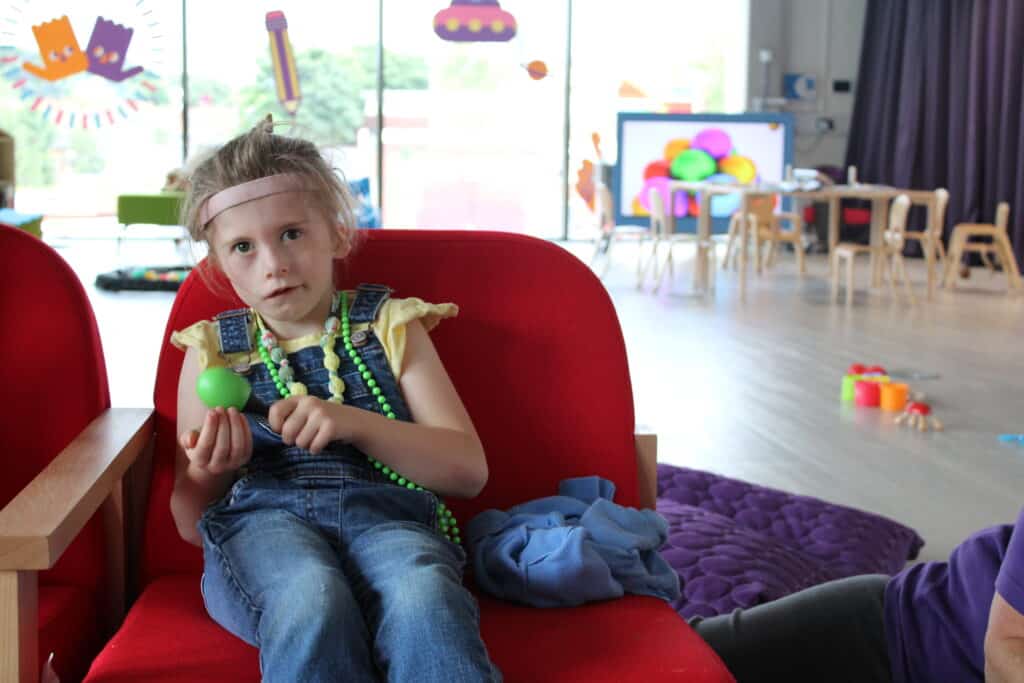 Charlotte, a young girl wearing dungarees, sitting in the Sense TouchBase Pears playroom holding a maraca.