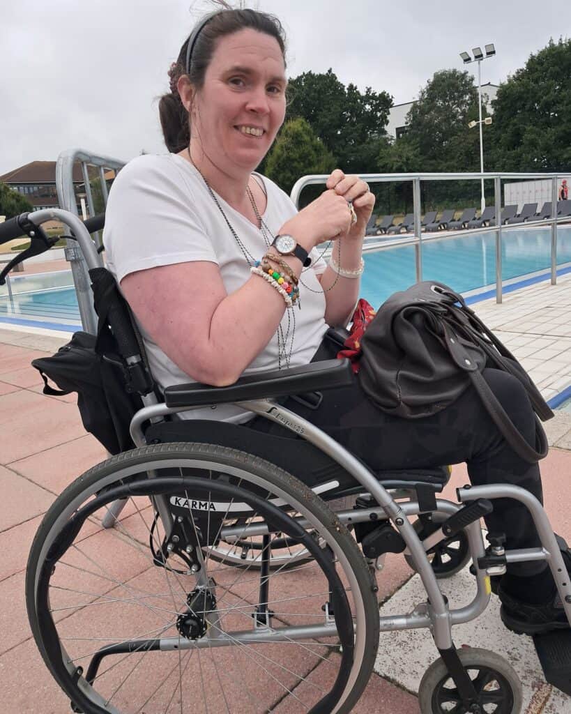 Joanna is sat in her wheelchair, in front of a swimming pool. She has lots of bracelets on her wrists, and is smiling back at the camera.