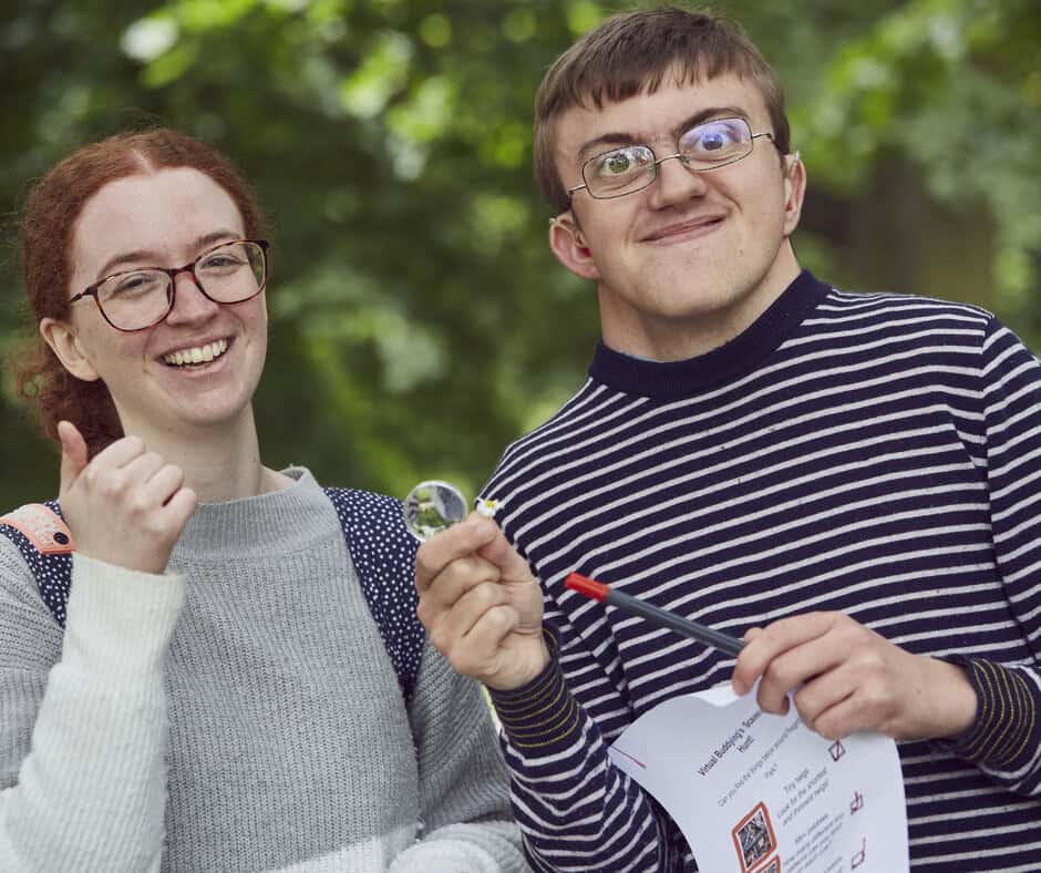A woman and a man standing and smiling in a park. She is giving a thumbs up and he is holding a flower.