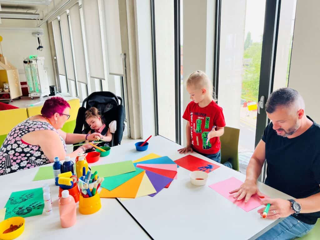 Verity, her support worker Anne, dad and sibling all playing at arts and crafts around a table.