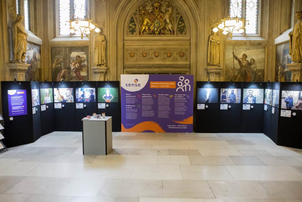 Exhibition stand in parliament. A large purple and orange board with information on surrounded by pictures of deafblind people and their stories.