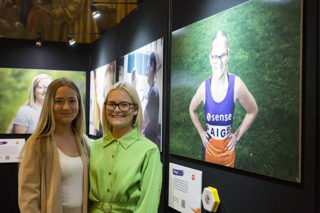 Two blond smiling women looking at the camera. They are stood next to a photo of one of the women in an exhibition