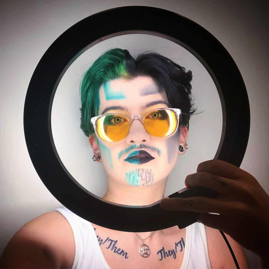An unsmiling face featuring white foundation, blue and grey highlights and tinted glasses holds a ring that is shining harsh white light onto their face. On their chest, they have 'they/them' tattooed twice.