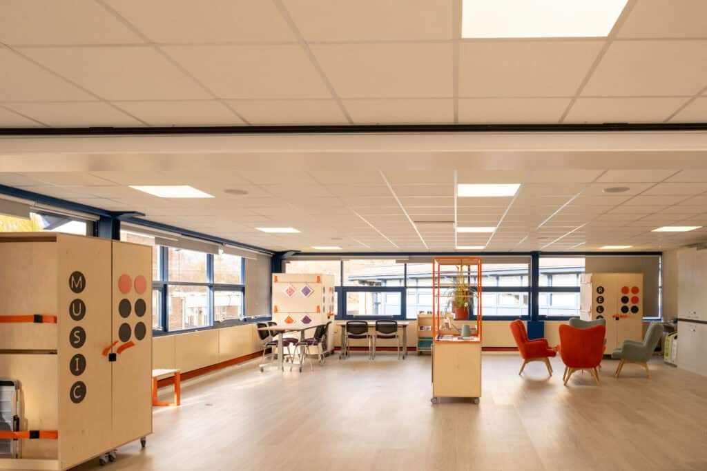 A large open room with lots of windows. In the middle of the room is a storage unite with an orange frame on top with hanging plants suspended. To the right of the unit are two green arm chairs and two orange arm chairs, arranged in a circle. To the left of the unit is two tables with chairs arranged around them. At the from of the photo is a bespoke storage unit with the word 'music' in black lettering.
