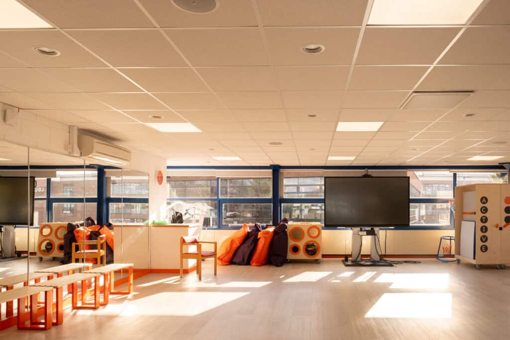 A large spacious room with lots of windows. On the left hand side of the room are floor to ceiling mirrors and a row of pale wood and orange benches in front. In the middle of the back wall is a large TV on a stand. To the left of the TV is a stack of purple and orange beanbags and a wooden storage unit. To the right of the TV is a wooden storage unit with 'active' in black lettering.