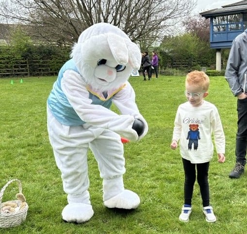 Alfie is stood to the right of a man in an Easter bunny suit. The bunny is holding out a ball for Alfie to play with. There is an Easter basket on the floor to the side of them.