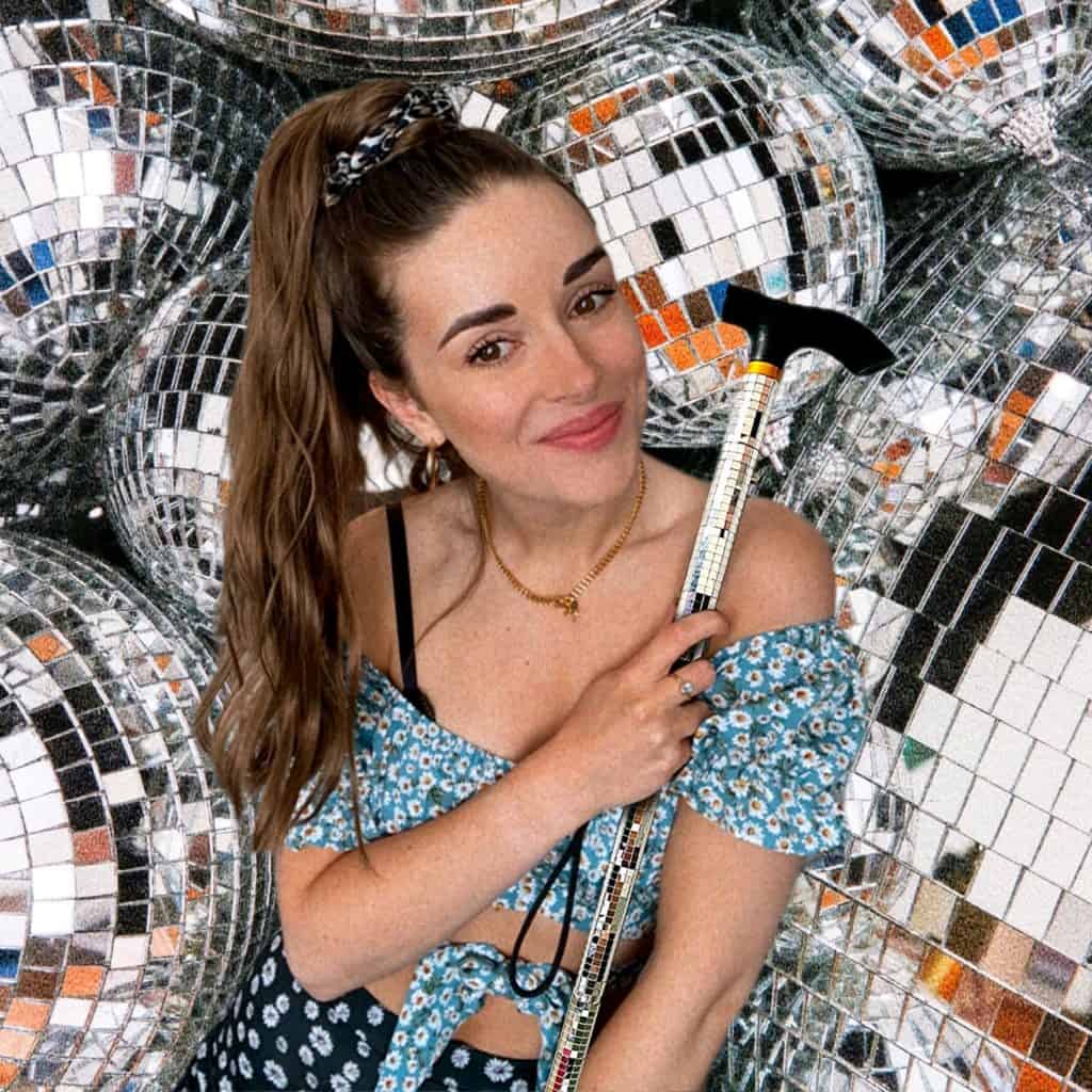 Alice Ella, a smiling white woman with brown hair in a ponytail, holds her mobility aid in front of a backdrop of disco balls.