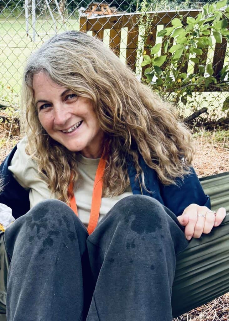 A smiling woman with shoulder-length blonde hair sitting on a green hammock outdoors
