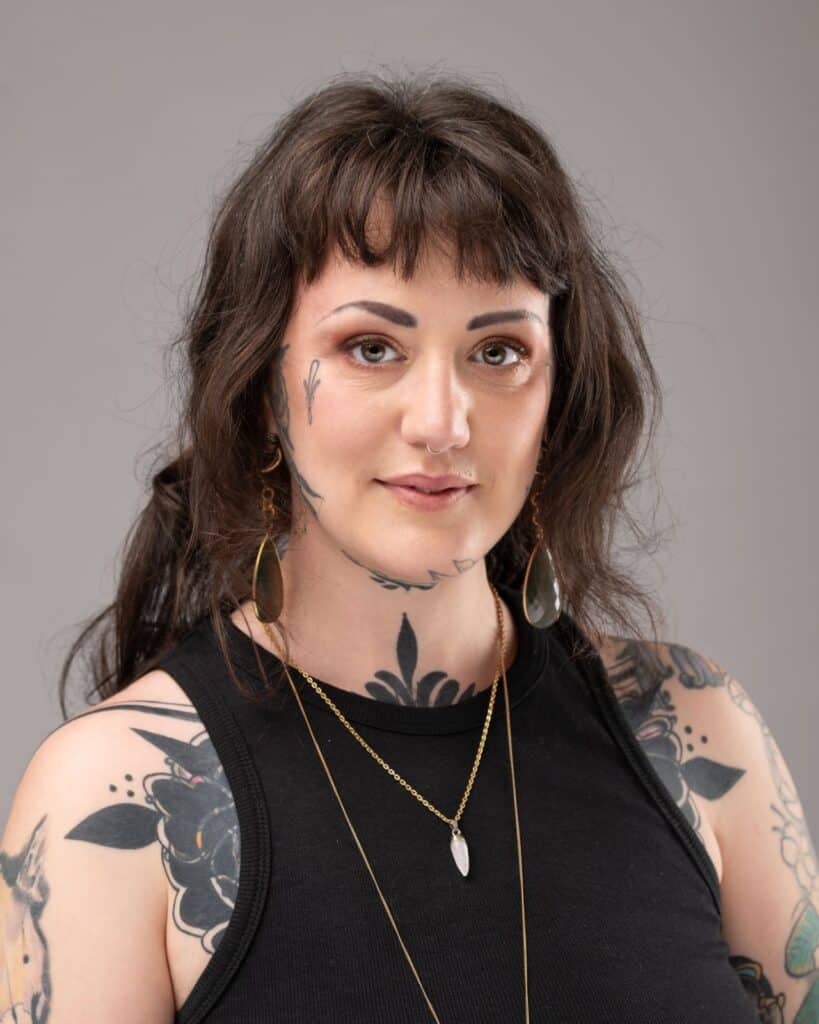 A woman looks at the camera. She has several tattoos, and is wearing large earrings.
