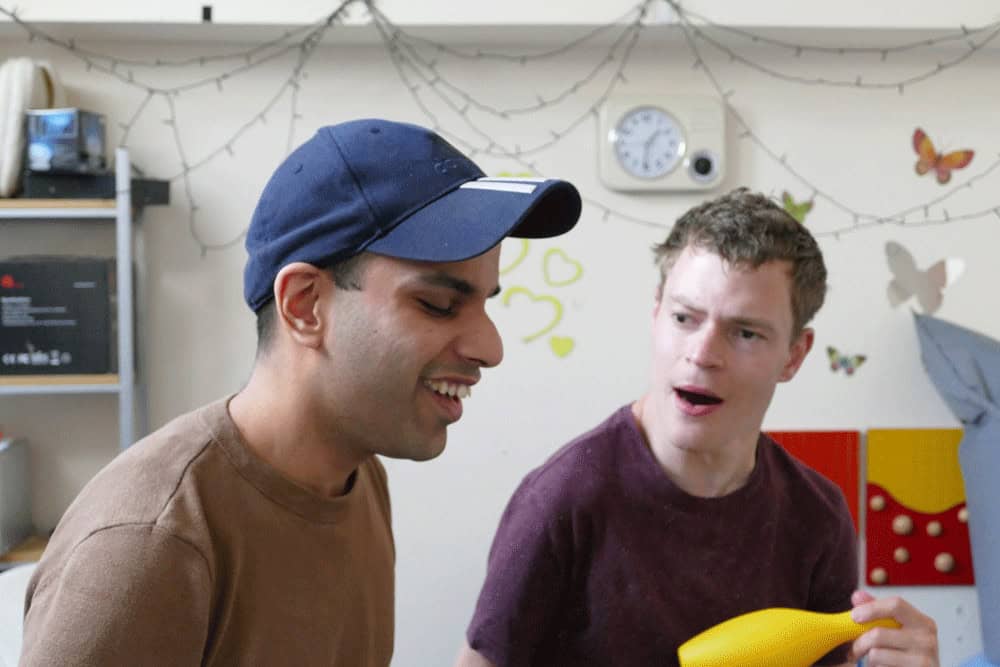 Two men sit near each other, one has his mouth open, the other is smiling