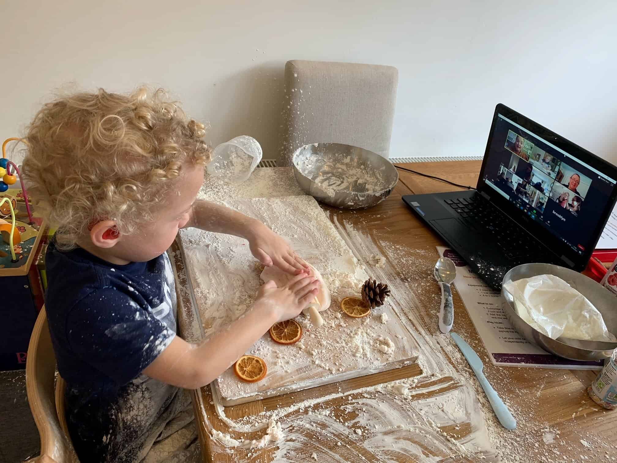 A small boy with blonde hair is rolling out dough infront of a Zoom call