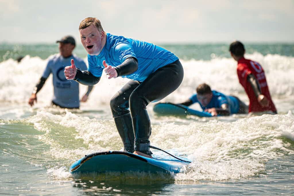 A young man in a wetsuit surfs on a calm sea, he's smiling and his thumbs are up.