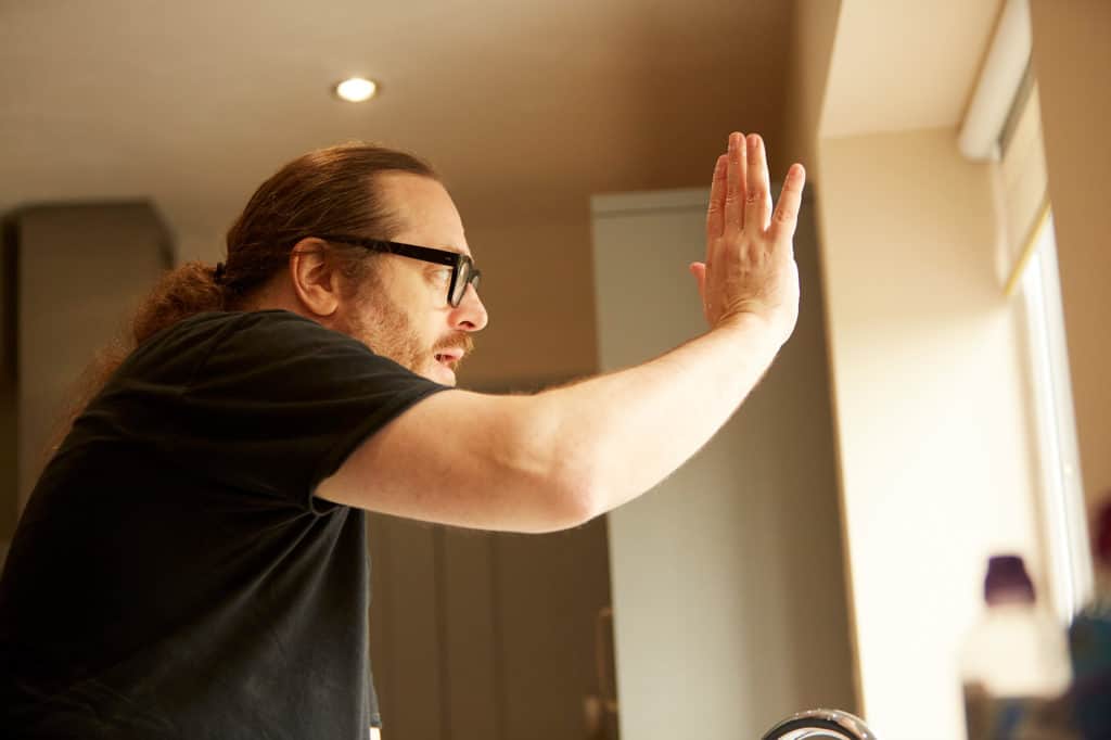 A man stands side on. He has long brown hair and glasses. He's wearing a black t-shirt and his arm is stretched out. It's Andrew. / --