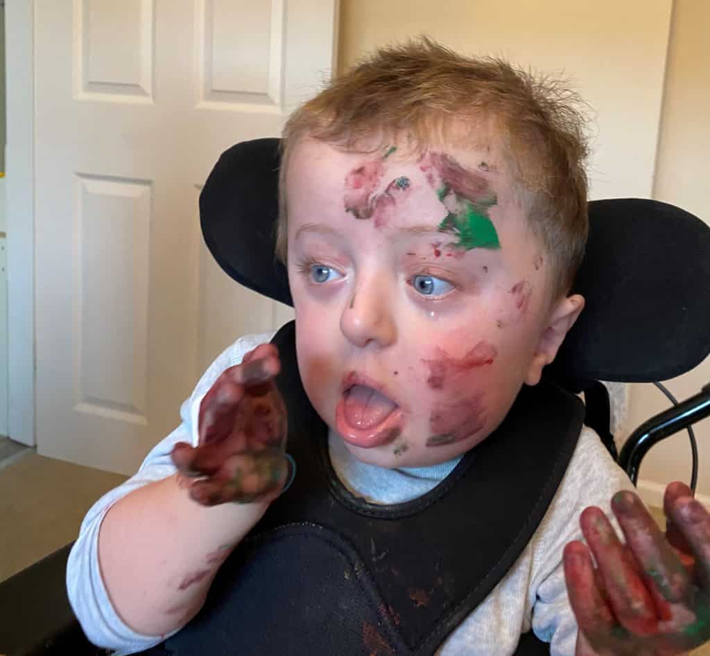 A little boy sitting in a wheelchair. He has brown and green paint on his hands and face. / --