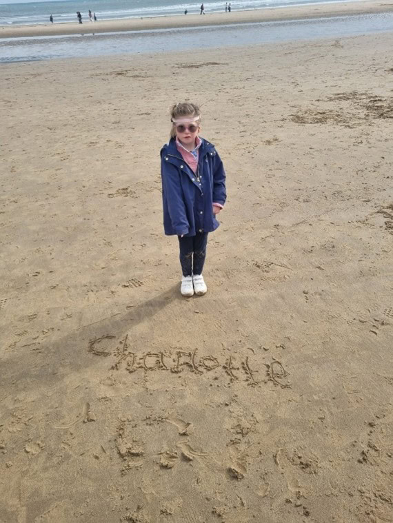 A young girl with sunglasses and a hearing aid head band stands on the beach. ‘Charlotte’ is written in the sand in front of her.