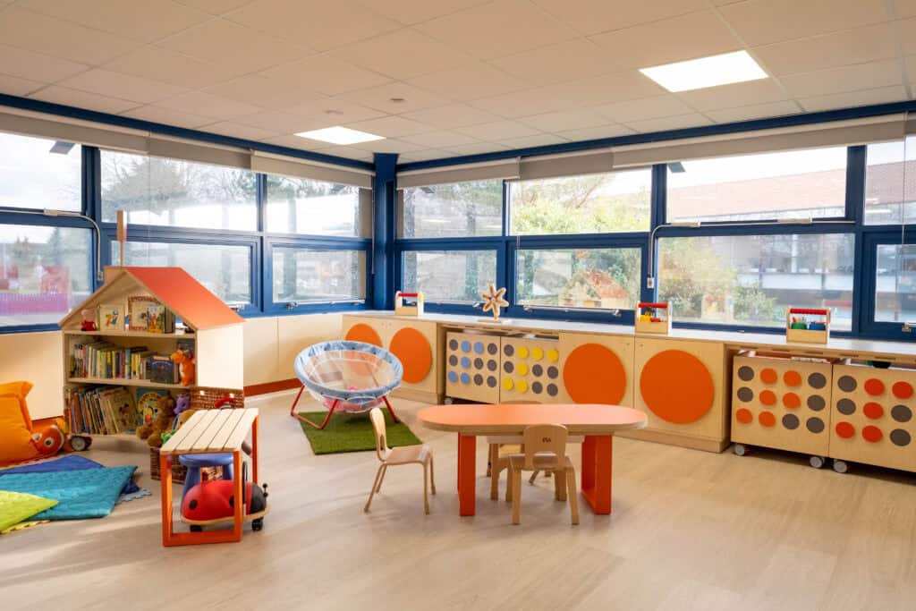 A large spacious room with windows covering all of the walls. There is a row of wooden storage units, covered in giant colourful braille dots along one wall. In front of the storage units is a small orange wooden table with pale wooden chairs, a storage unit shaped like a house filled with booked and colourful mats on the floor.