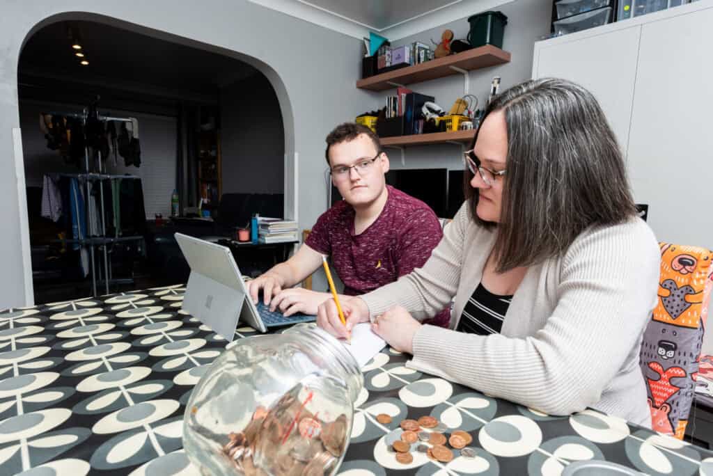 At a table, a woman with short dark hair and glasses is writing on a notepad. Next to her, a young man with short dark hair and glasses is looking concerned and typing into a computer.