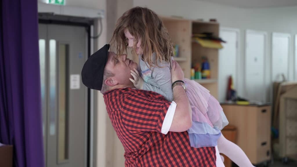 A man in a red shirt holds his young daughter in his arms, she reaches out to touch his face.