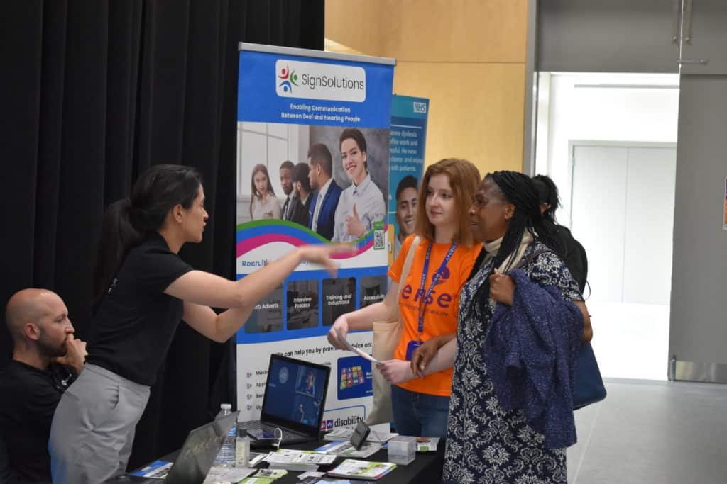 A woman with braids, linking arms with a woman wearing a Sense t-shirt, talks to a woman at a stall at the Pan Disability Jobs Fair.