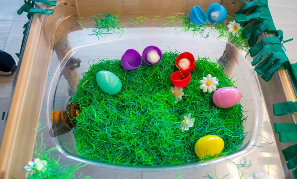 A tray filled with green string, to represent grass, plastic flowers and eggs.