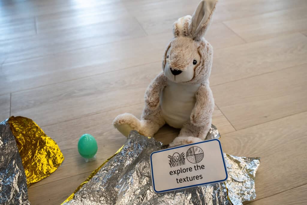 A furry bunny toy is sat on gold foil. There is a picture symbol card, leaning against the bunny, that reads 'explore the textures'.