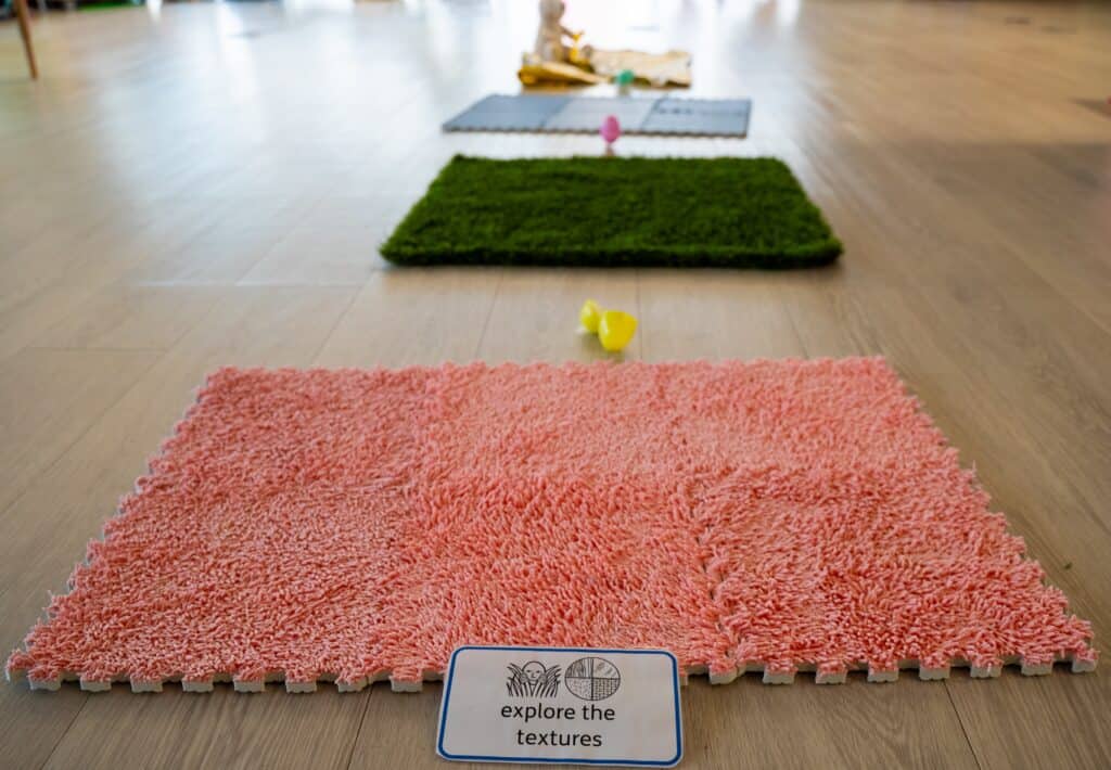 A card at the bottom of the picture reads 'explore the textures'. It is leaning against a carpet square, with a grass square directly after it and a plastic square after that one. There is a toy bunny at the end of the path.