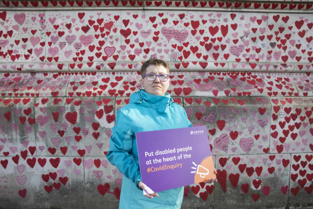 Landscape shot of the National Covid Memorial Wall covered in hand drawn red and pink hearts. A woman in a blue coat and glasses stands in front, facing the camera. She is holding a purple and orange sign reading 'Put disabled people at the heart of the #CovidInquiry.'