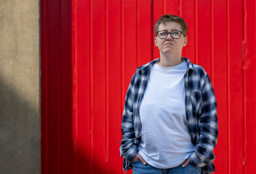 A woman stands in front of painted doors. She has her hands in her pockets and is frowning.