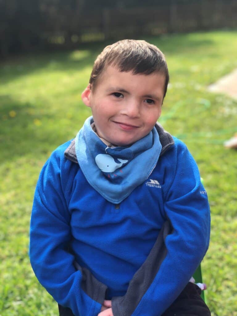 A young boy in a blue shirt and bandana sits in a garden.