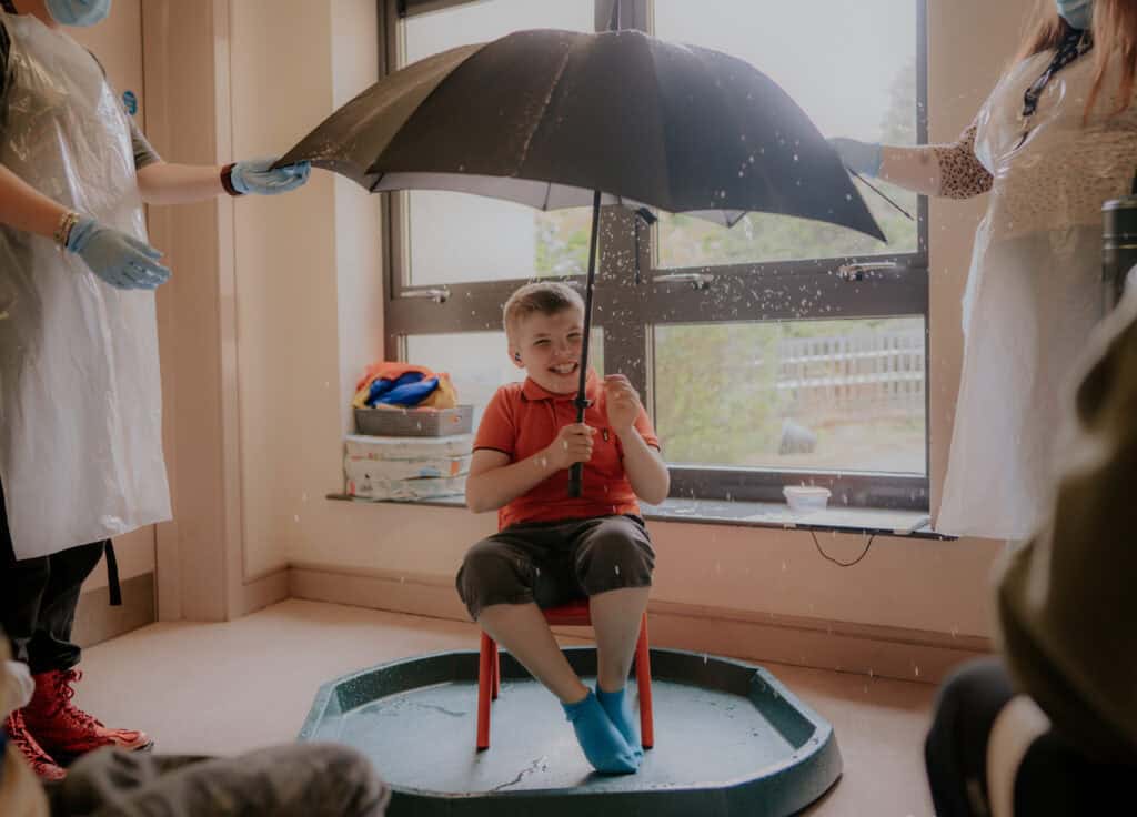 A smiling young boy sits inside under an umbrella with water spilling off it.