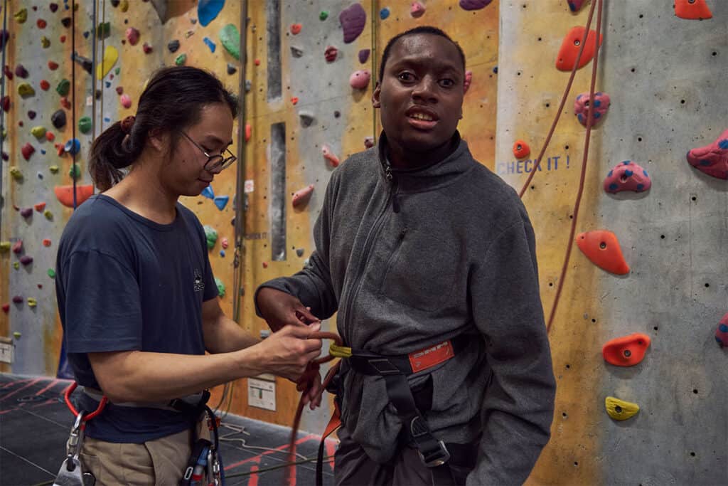 Franklyn is wearing a grey fleece and a harness which is being secured by a helper. A climbing wall is in the background.