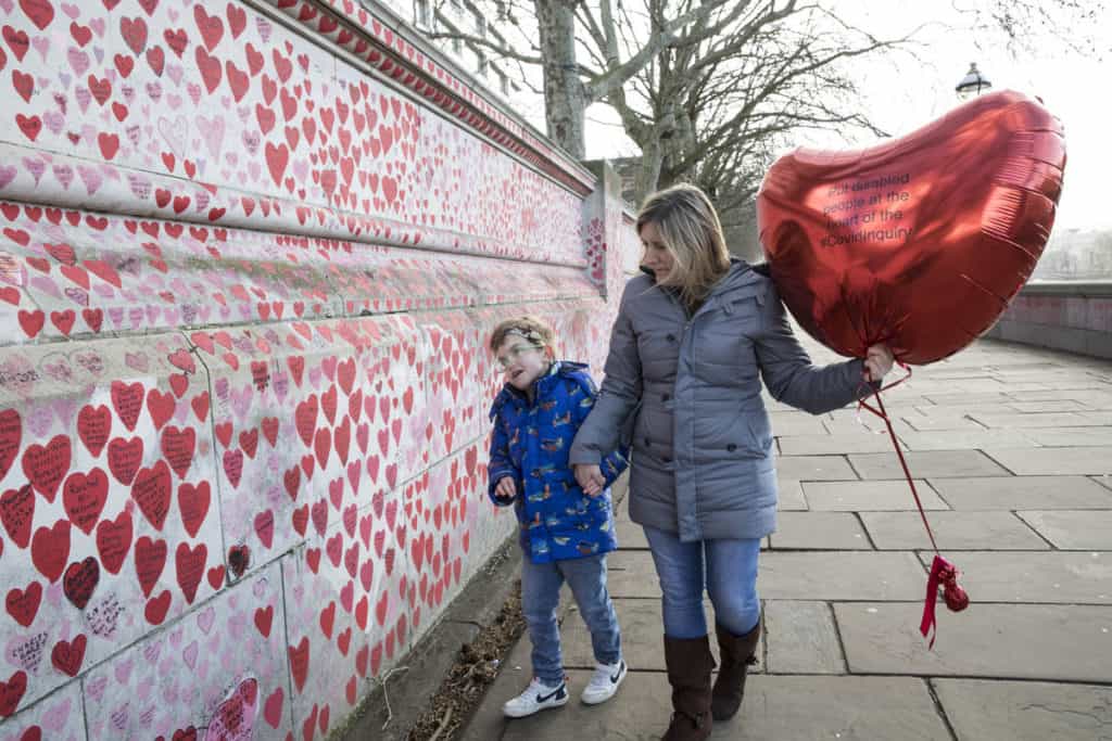 A woman and her son walk alongside the Covid-19 memorial wall, covered in red hearts. They're holding a big, heart-shaped balloon with "Put disabled people at the heart of the Covid inquiry" written on it.
