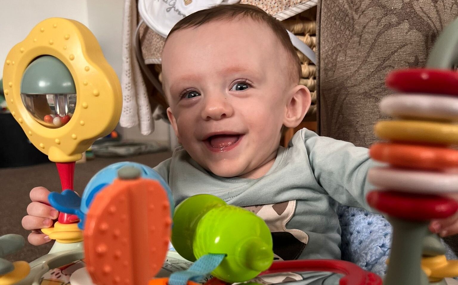 Gracen is sat in a high chair, smiling widely at the camera with his bright eyes. He is holding a set of multi coloured toys in front of him.
