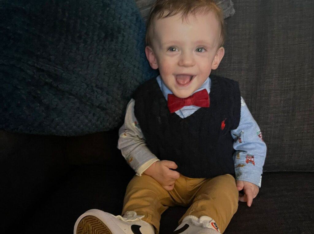 Gracen is sat on a sofa smiling with his mouth wide open and tongue on show. He is dressed smart in a shirt, with a red bow tie, navy jumper and brown trousers.