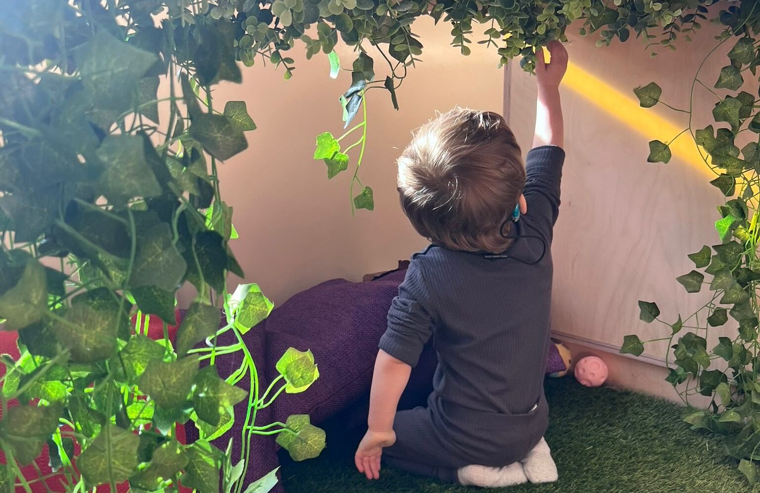Gracen is kneeling inside of a wooden sensory den. The sensory den has vines hanging from the roof, which Sonny is reaching up to feel.