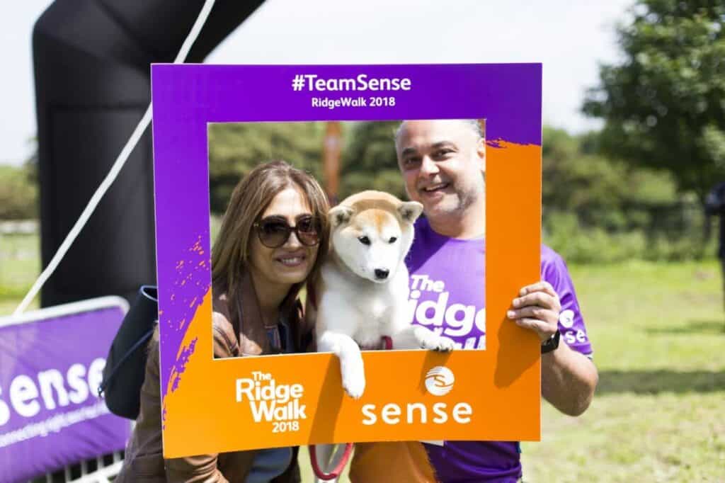 Guni Suri and his Shiba Inu, Stella, and a woman wearing sunglasses pose in a Sense photo frame at the end of the RidgeWalk Challenge.