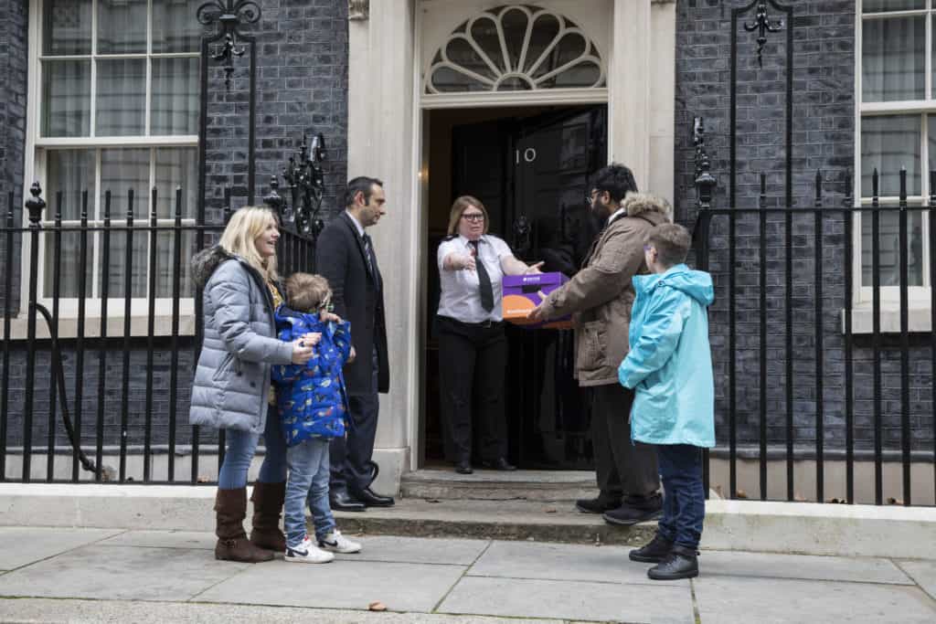 Landscape shot of four adults and one child standing on the steps in front of Number 10, Parliament. One of the group hands purple box to a female police officer who is standing in front of the open door.