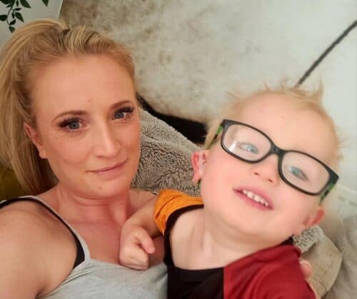 A young boy wearing glasses smiles at the camera next to his mum, who has blonde hair and is wearing a grey t-shirt